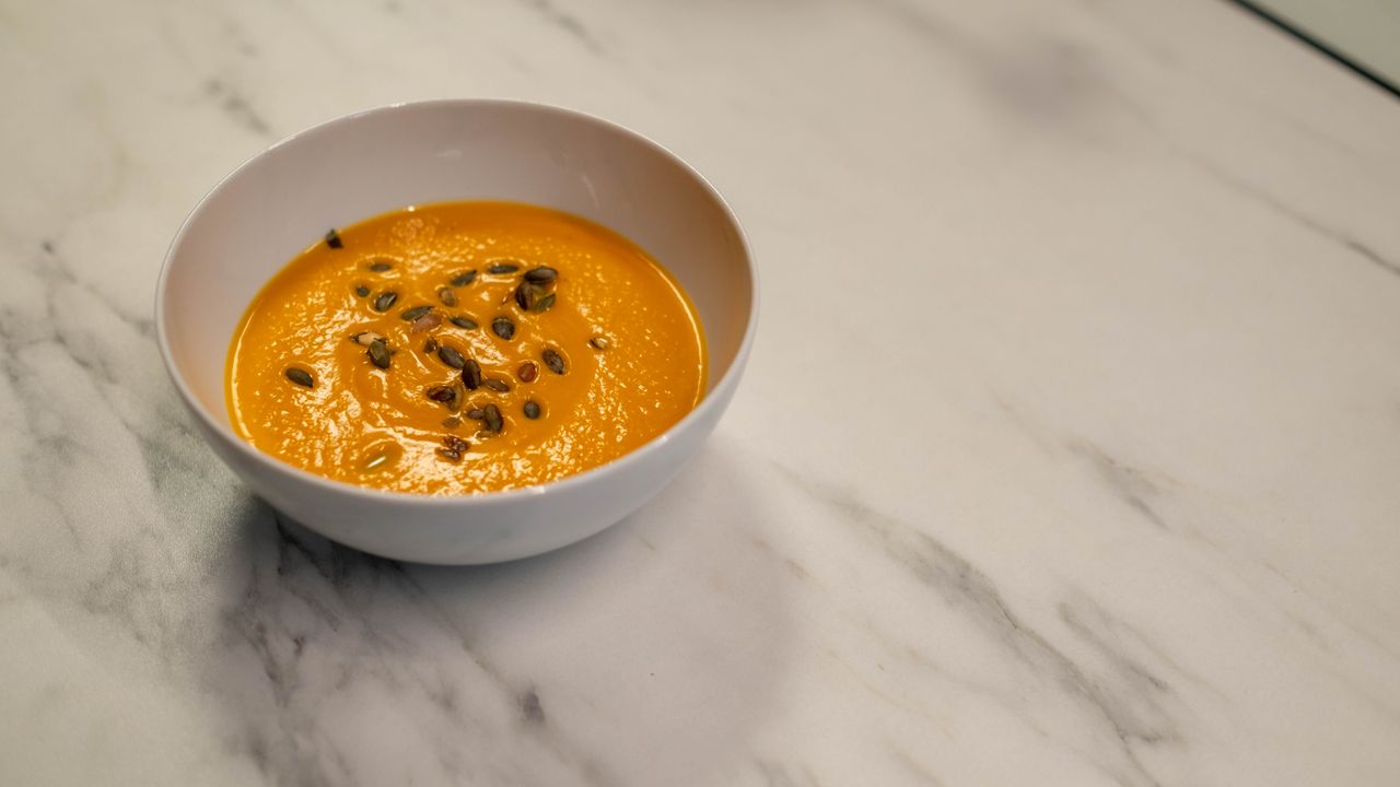 Bowl of creamy orange pumpkin soup topped with toasted seeds on a white marble counter