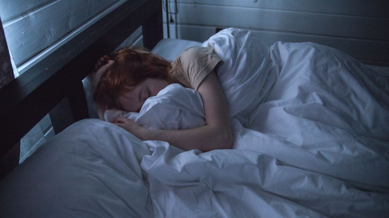 Woman with red hair sleeping peacefully under white sheets in a dim bedroom