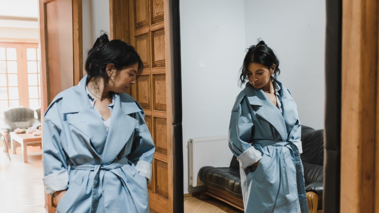 Woman in a pale blue trench coat looking thoughtfully at her own reflection in a tall wooden framed mirror
