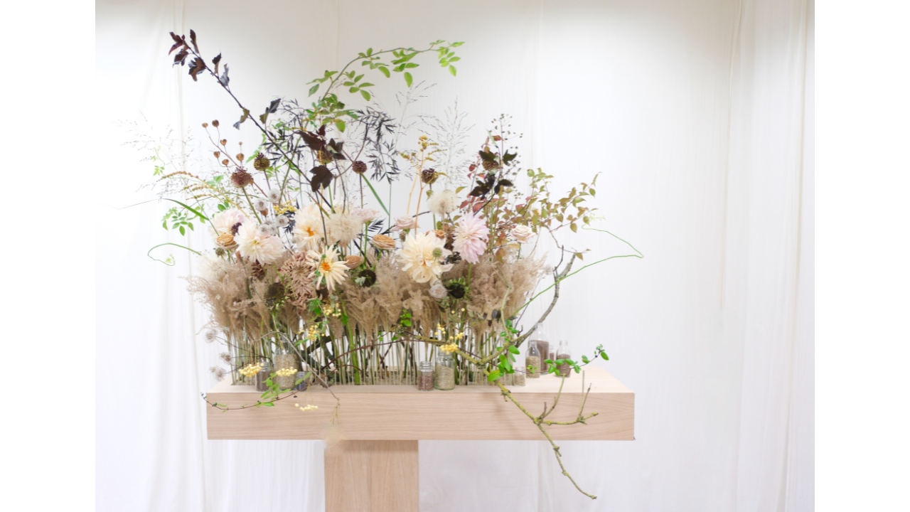 Large-scale lobby floral installation by Joseph Massie, winner of UK Florist of the Year 2024 at BFA FleurEx, featuring pale dahlias, grasses, seed heads and seasonal foliage arranged in naturalistic layers on a wooden plinth for a Nottingham chemist headquarters.
