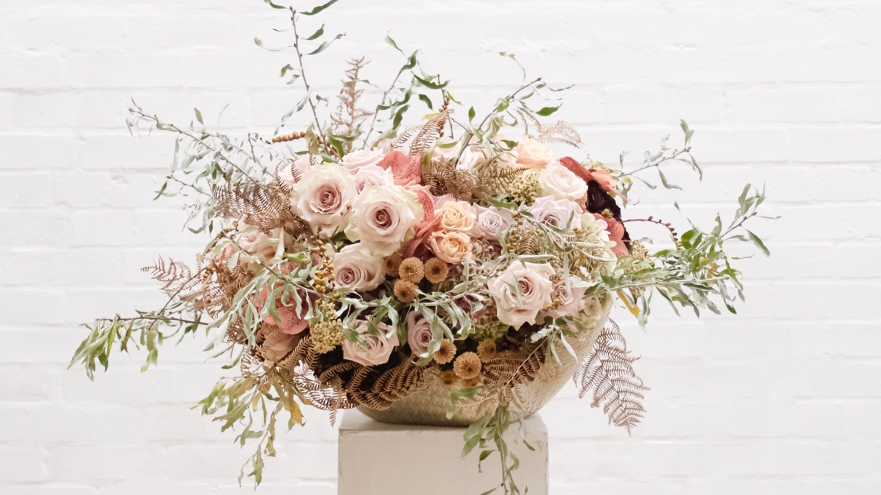Elegant fan-shaped floral arrangement featuring blush pink roses, lilac blossoms, deep plum ranunculus, and dark hellebores in a sculptural black metal vase. Contemporary floral design by Joseph Massie.