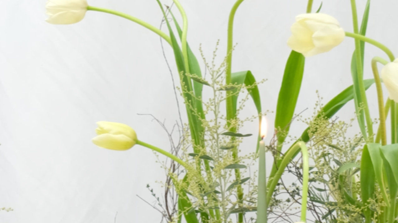 Minimalist spring floral arrangement featuring soft yellow tulips, delicate greenery, and textured moss, styled with a muted color palette on an elegant tablescape with ceramic plates and a lit tapered candle.