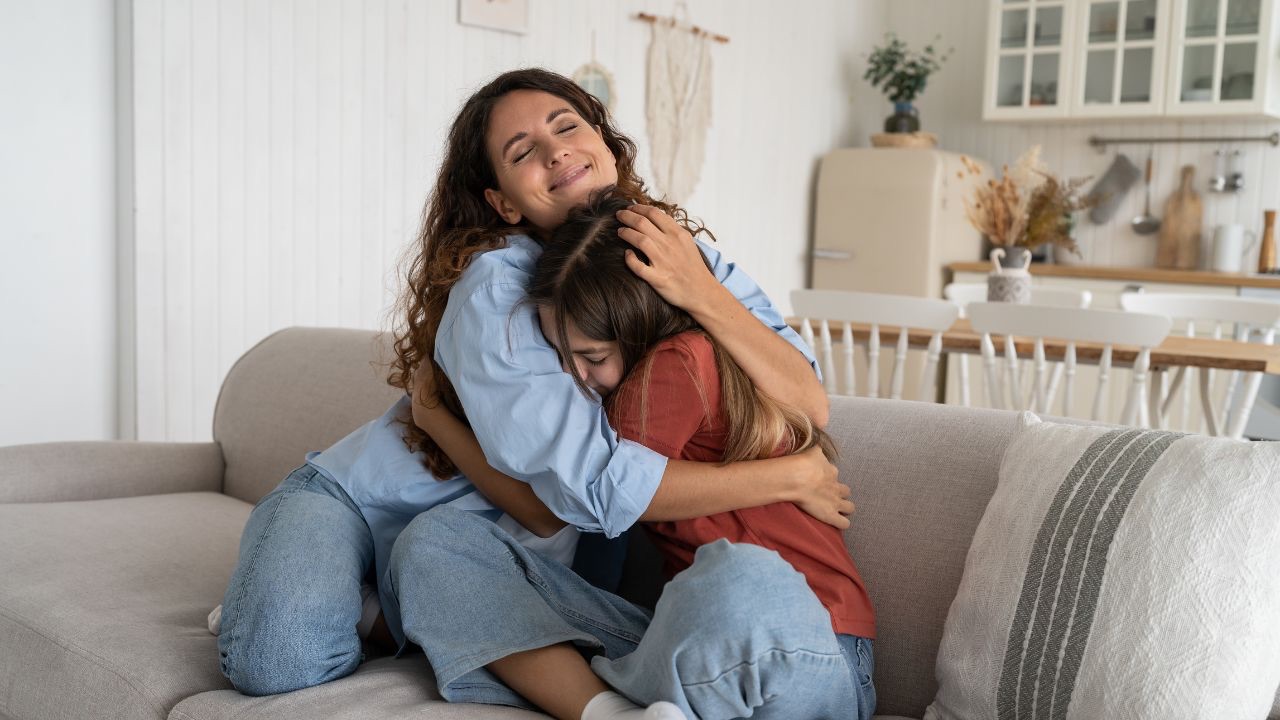 Image is of a parent hugging a child, on the couch. The parent is wearing a denim long sleeve top, and light colored jeans, has long curly hair, and is smiling with their eyes closed. The child is wearing a dark pink long sleeve top and light colored jeans, and is leaning in toward the parent with their eyes closed and a smile on their face. Parenting a transgender child has taught me that love is the easiest gift I can give my kids. 