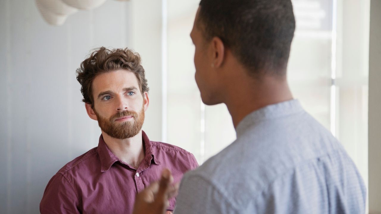 Two people are standing facing each other, talking. One has short hair and a beard, and is wearing a raspberry colored, colored button down shirt. The other is has buzzed, dark hair and skin, and is wearing a light blue dress shirt. It can be difficult to tell others that our children are transgender, but so long as you have the support of your child and you are protecting their safety, you can tell people that you trust. 