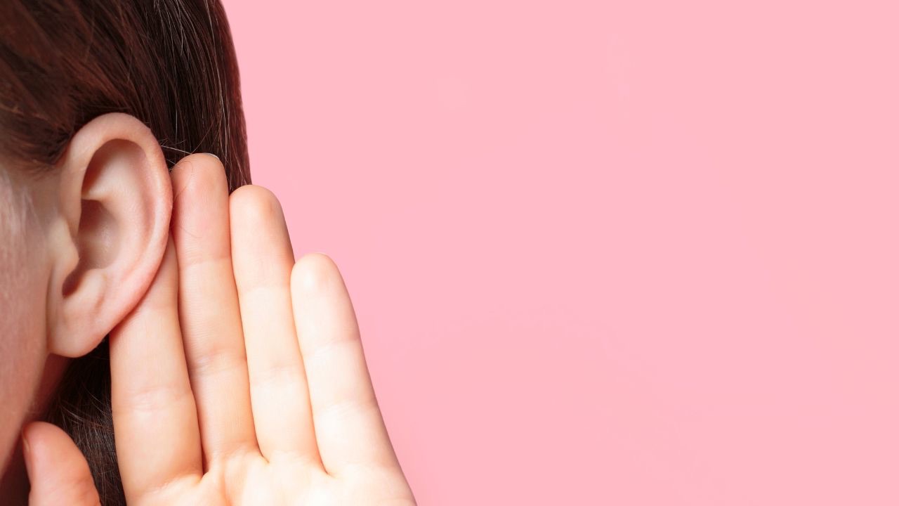 Image is the side of a person's head from the ear over, with a hand being held up to the ear, against a pink background. We need to listen better to what our children are saying to us, especially when they are telling us what it means for them to be transgender. 