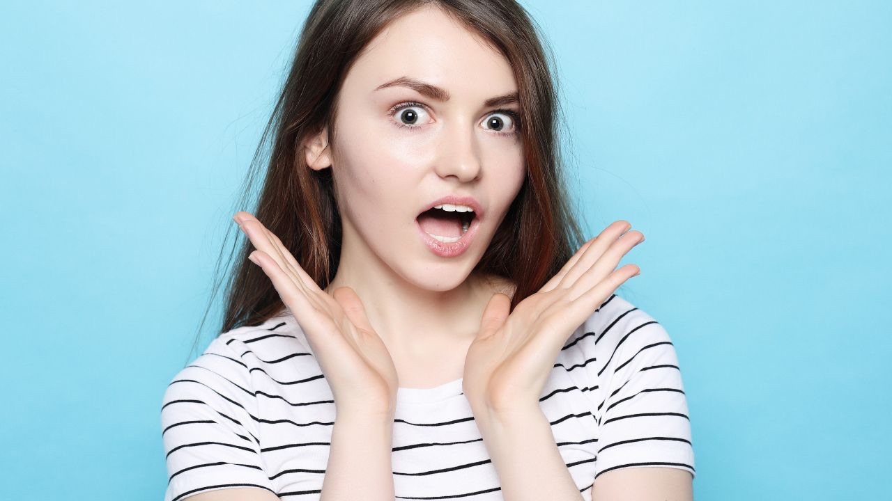 Image is of a person with big eyes and a mouth open wide, in a white shirt with their hands wide open under their chin and a surprised look on their face, in front of a blue background. Life doesn't always look how we expect, especially when our children come out as transgender and the life we thought we were living doesn't look like the one we planned. 