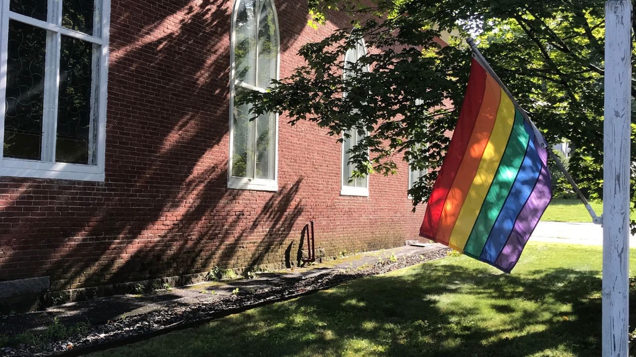 Image is of a brick church building with a white sign pole that has a rainbow flag hanging off it. It's not enough to hang a flag out to show you welcome everyone. You need to know if the church is Open and Affirming. 