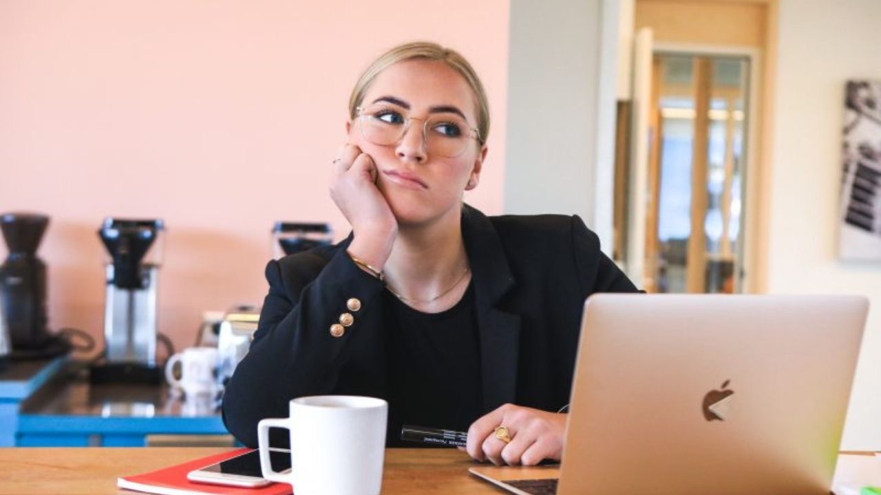 Image is a person in a black shirt sitting in a pink kitchen at a counter or table, in front of a laptop with some notebooks and a cup of coffee. Their head is propped on their hand and they have a confused look on their face. You don't have to have everything figured out when your child comes out as transgender. 