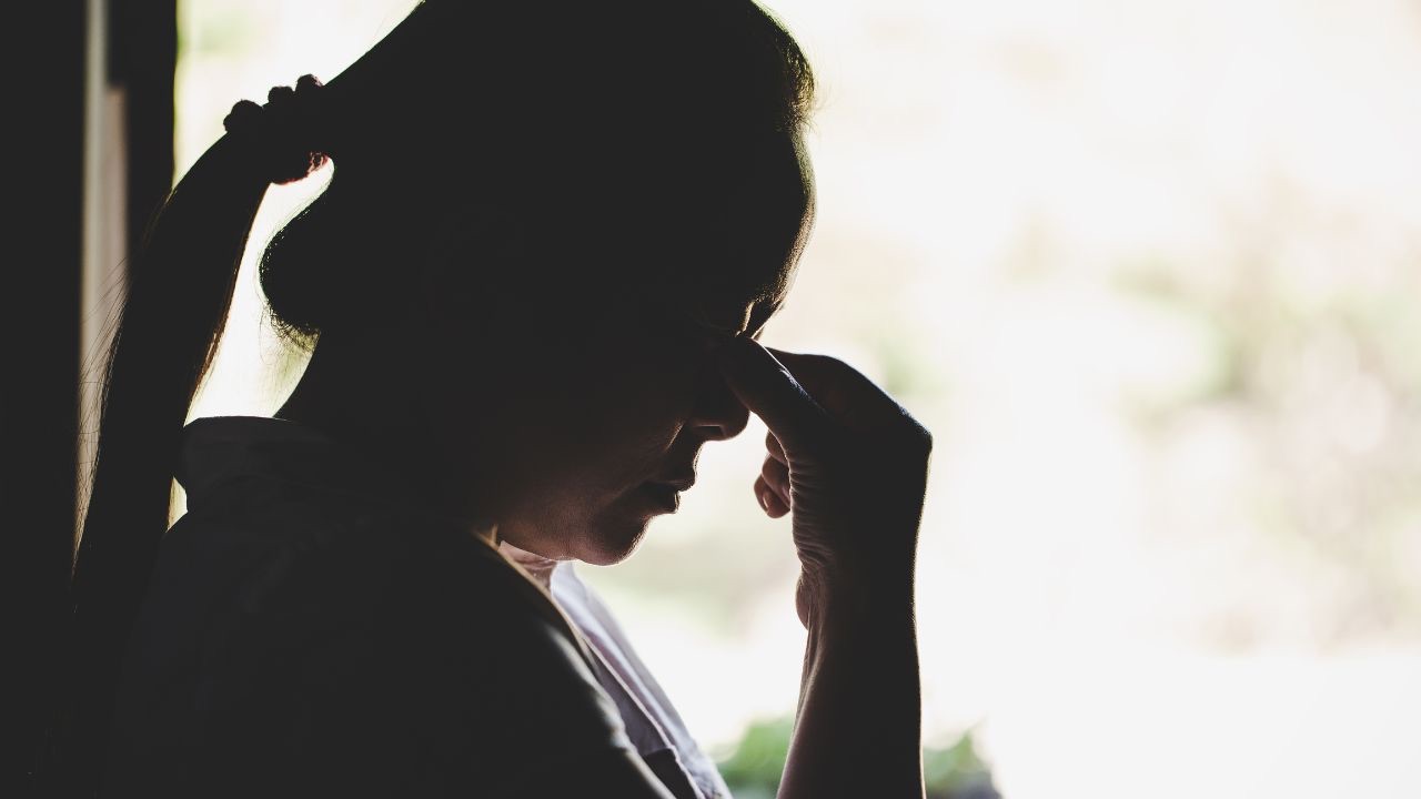 Image is a woman's silhouette, standing in front of a window. She has her head down and her hand up to her eyes. Because dealing with unsupportive people when our children come out as transgender is frustrating. 