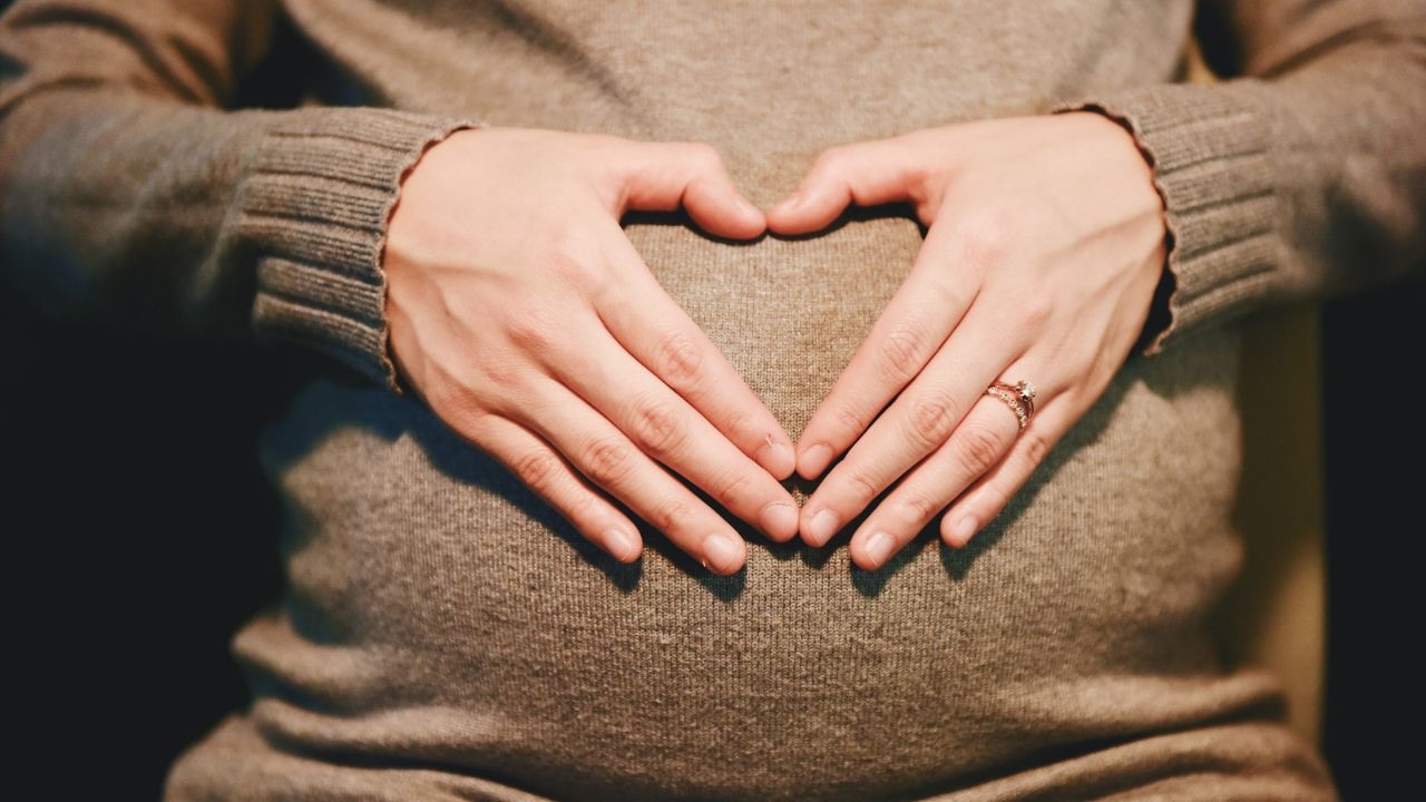 Image is of the belly of a pregnant person in a brown sweater, and their hands making a heart over the belly. When I was first pregnant with my oldest I chose a list of names that would later be used for both of my children. Neither of those names are used by my children now, something I sometimes struggle with. 