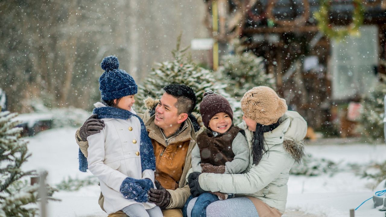 Image is a family of four crouched down outside in the snow. The parents are each smiling at one of the kids. A few small changes are all it takes to make the holidays feel affirming for your transgender child. 