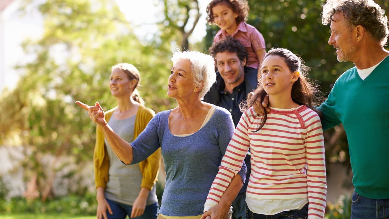 Image is of a group of extended family members walking together outside, arms jointed, a young child on the back of an older family member. They are all smiling and one of the eldest family member is pointing ahead to something in the distance. Getting family on board with your transgender child can take time, hard conversations, and boundaries, but it's not impossible. 