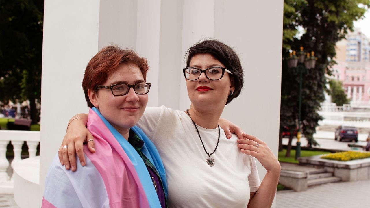 Image is two people standing in front of a post. One has the transgender pride flag wrapped around their shoulders, and the other is wearing a white tee shirt and a necklace. Both are smiling and looking toward the camera. During transgender awareness week, it's important to listen and learn from the voices of transgender people.