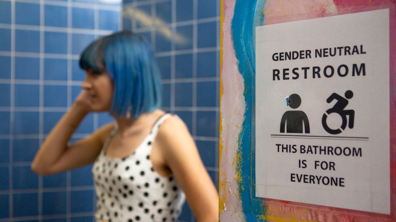 Image is of a person in a white tank top with black dots, with blue hair holding a phone to their ear in front of a blue tile wall. On the wall next to them is a sign that reads, "Gender Neutral Restroom. This bathroom is for everyone." In the sign is a photo of a person in a wheel chair and a person with no gender. 