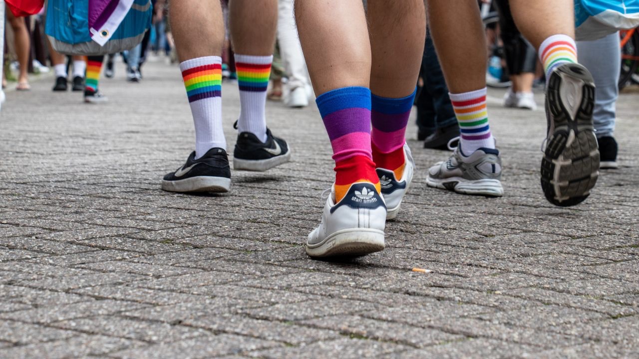 Image is a bunch of legs, all wearing socks with different LGBTQ flag colors on them, walking in a parade. Because there is strength in the LGBTQ community. 