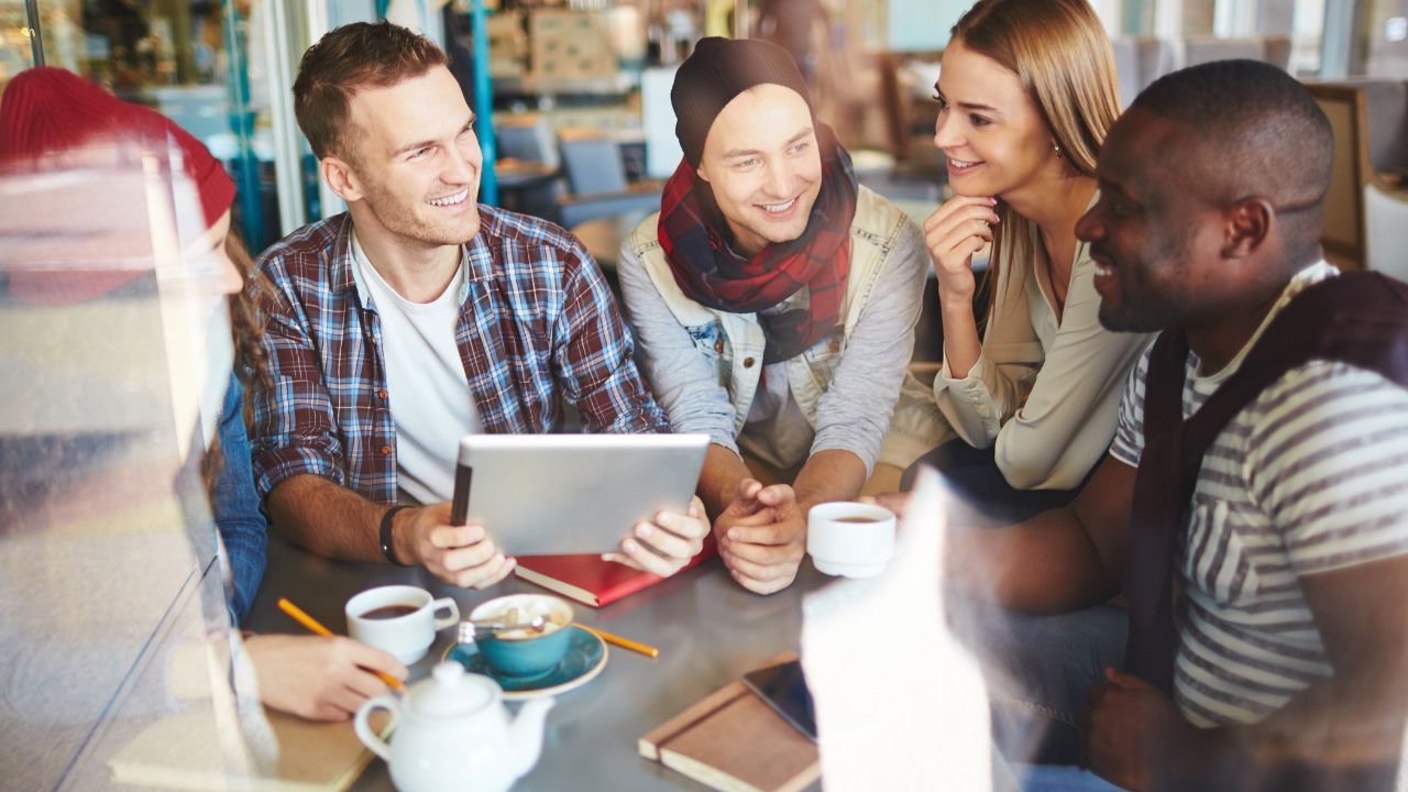 Image is a group of people of all races and genders sitting around a table in conversation, smiling. A big part of transgender awareness is having a strong community to support you after coming out. 