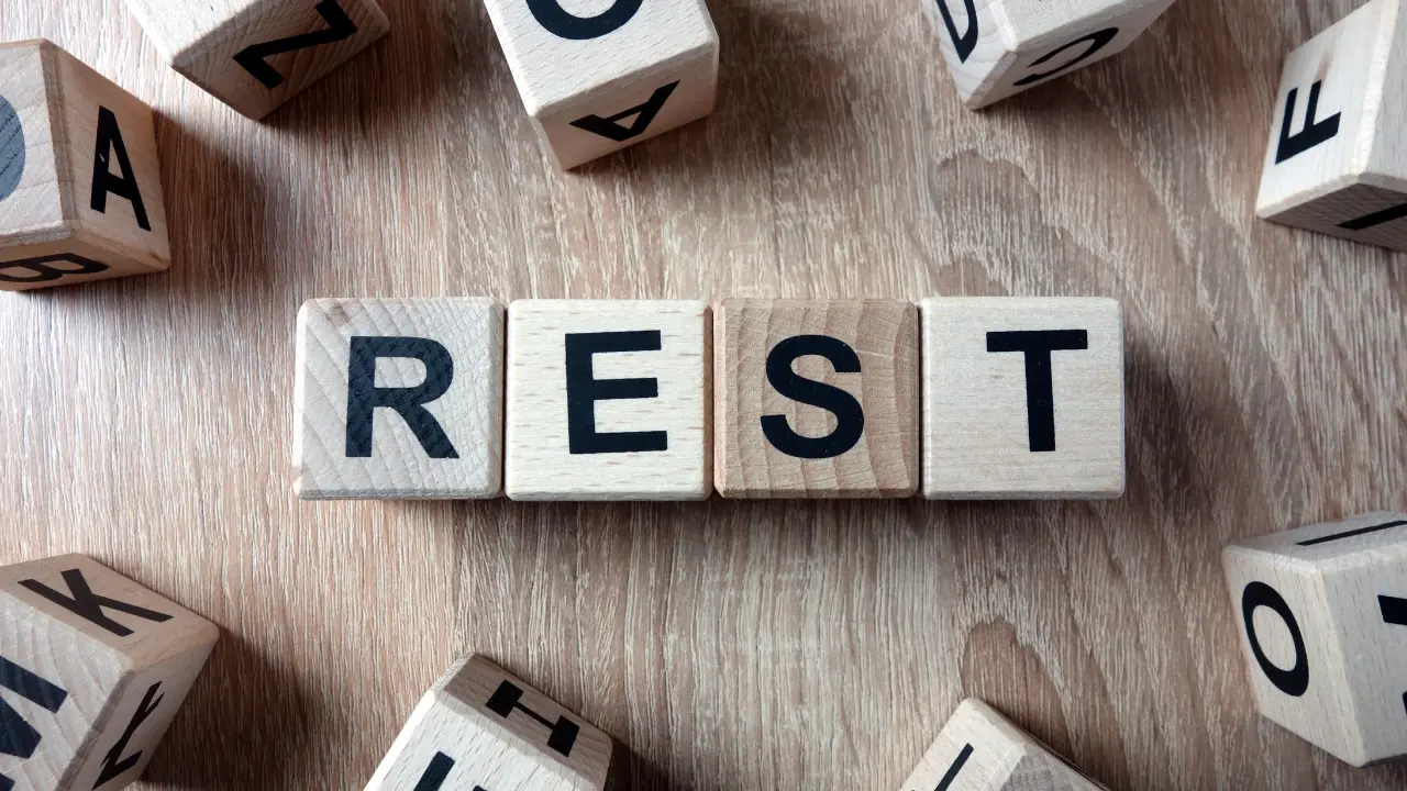 Wooden blocks spelling the word REST centered among scattered letter cubes on a wooden table.