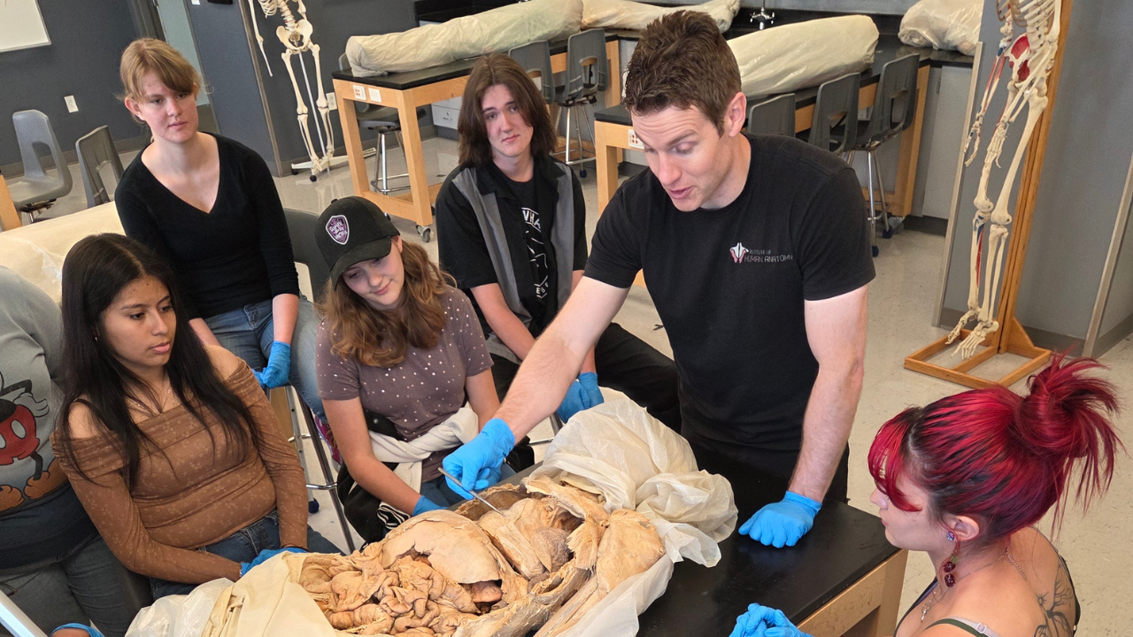 Students wearing blue gloves observe a hands-on human anatomy lesson and dissection session inside a university science lab.