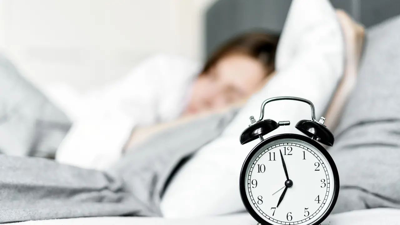 A black vintage alarm clock showing 7:00 on a nightstand with a blurred woman sleeping in bed in the background.
