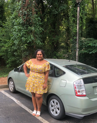 Nancy standing in front of a car, with a background of woods, wearing a sunny yellow dress