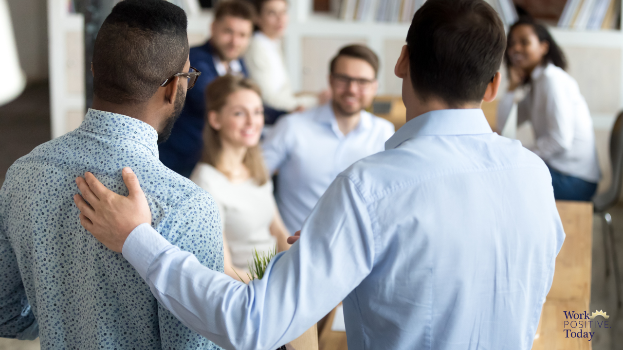 Leader or mentor in white shirt with welcoming hand on shoulder of new employee in patterned blue shirt, introducing him to smiling team members in bright modern office, demonstrating effective onboarding buddy system that increases retention 50% and helps new hires grow roots in company culture from day one, with Work Positive Today logo visible.