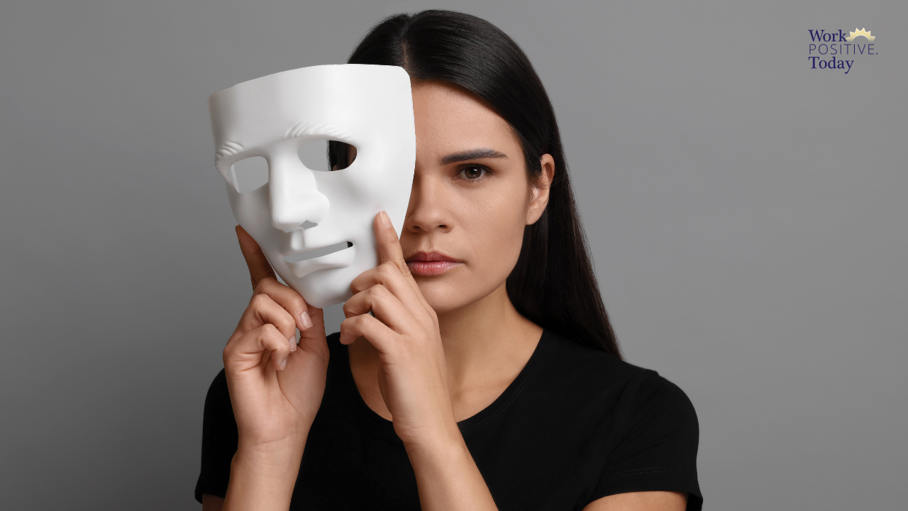 Professional woman in black shirt holding white mask partially away from her face against gray background, revealing her authentic self on one side while showing the facade on the other, illustrating the shift from pretending perfection to authentic leadership that creates psychological safety where 84% of employees want to learn from mistakes, with Work Positive Today logo visible.