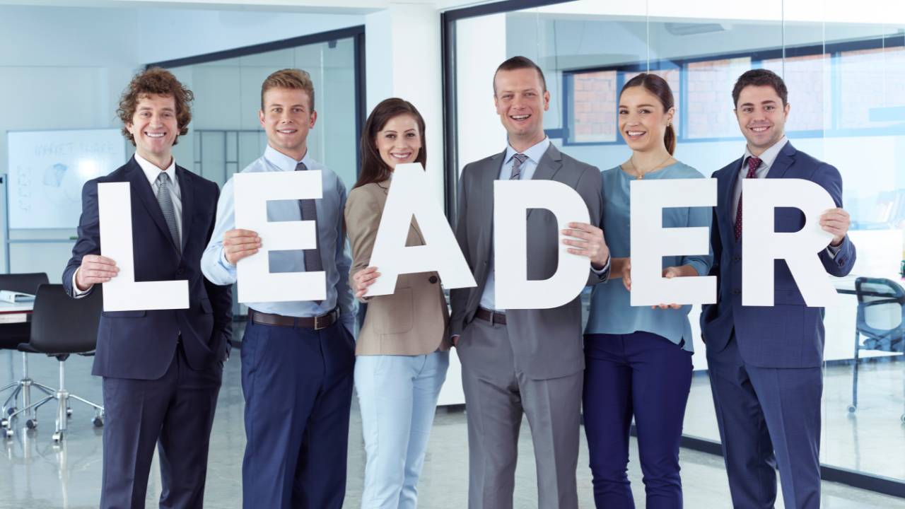 Six diverse smiling business professionals in suits standing in modern office holding white letters spelling 