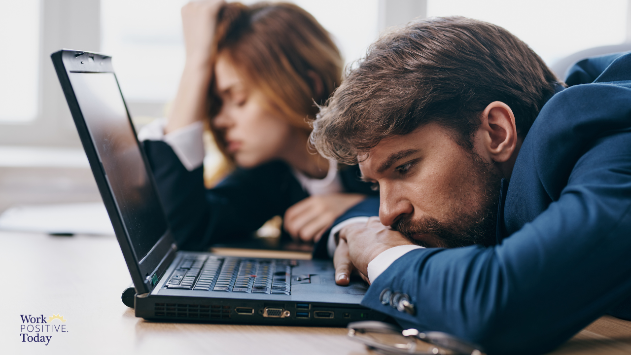 Discouraged businessman in blue suit with head resting on hand looking down at laptop with female colleague blurred in background, both appearing disconnected and isolated despite working together, illustrating workplace loneliness epidemic affecting 86% of workers, with Work Positive Today logo in corner.