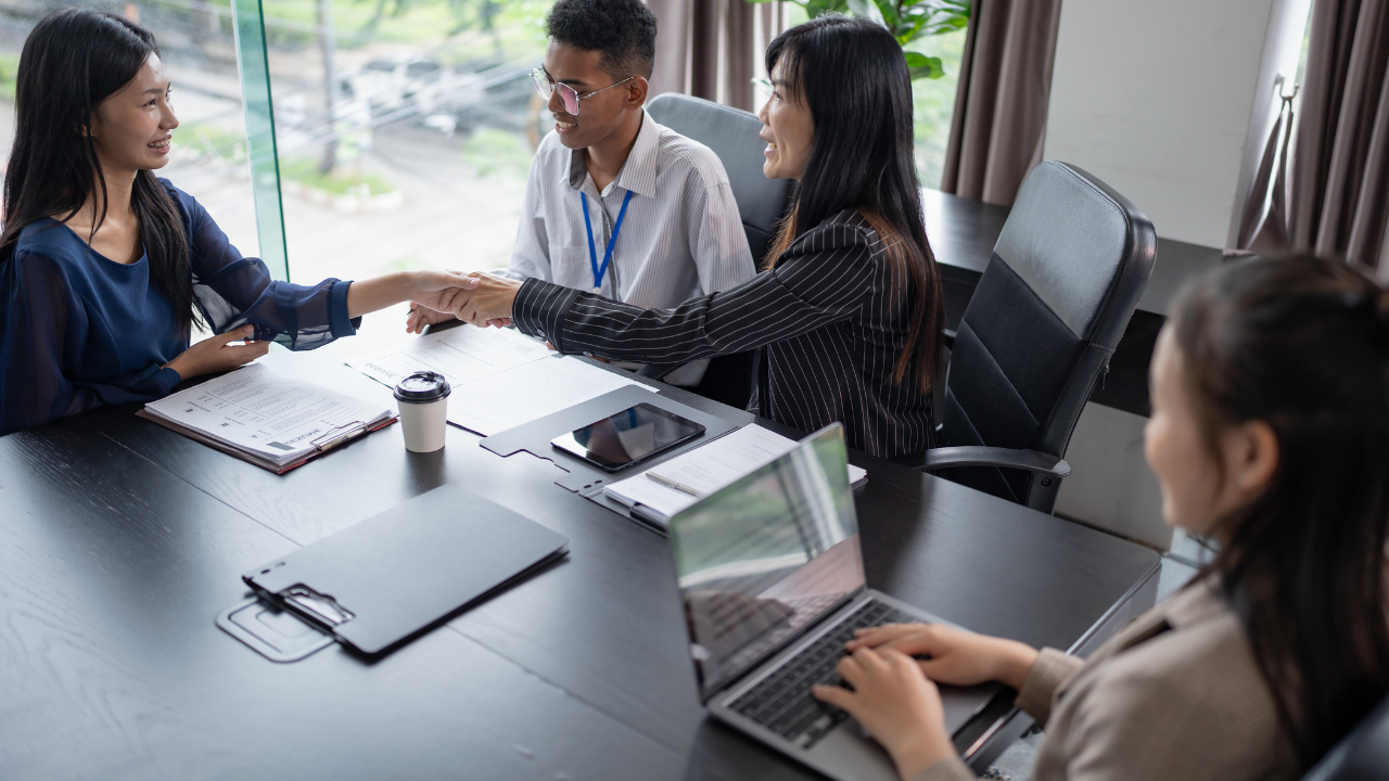 Three business professionals shaking hands across conference table while fourth person works on laptop, with documents and coffee cup on table in bright modern office, representing successful talent attraction, cultural fit hiring, and positive workplace culture that retains top employees.