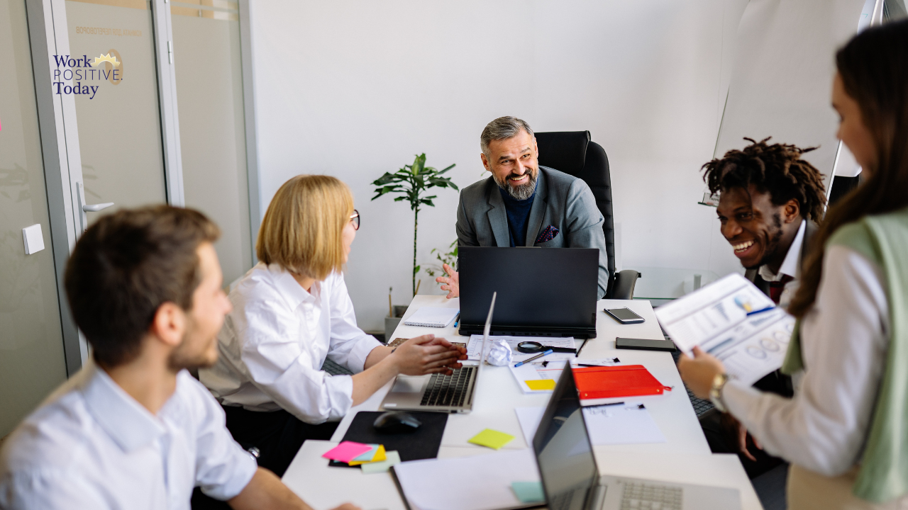 Diverse team of five professionals in bright modern office engaged in collaborative meeting with genuinely happy expressions, smiling leader in gray suit facilitating meaningful discussion, demonstrating authentic workplace joy and fulfillment through purpose-driven work and genuine relationships rather than superficial perks, with Work Positive Today logo visible.