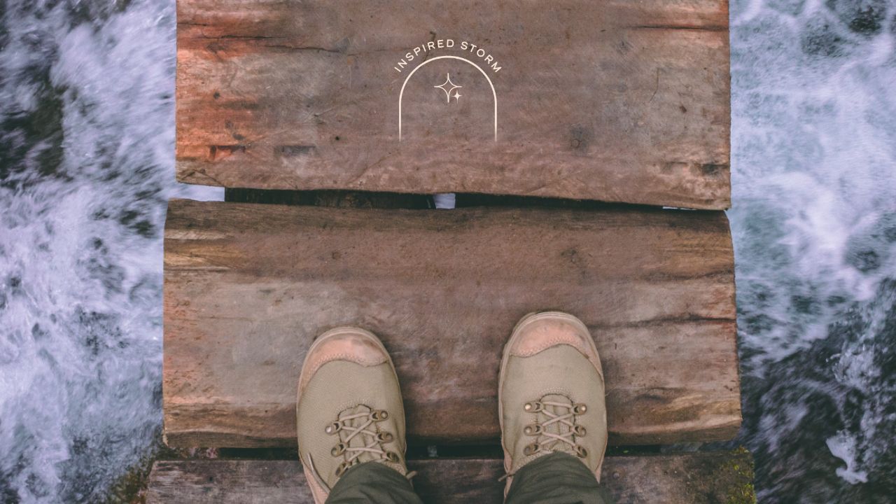 Picture is a personal point of view looking down at their hiking boots. They are standing on a wooden bridge floating over a raging river.