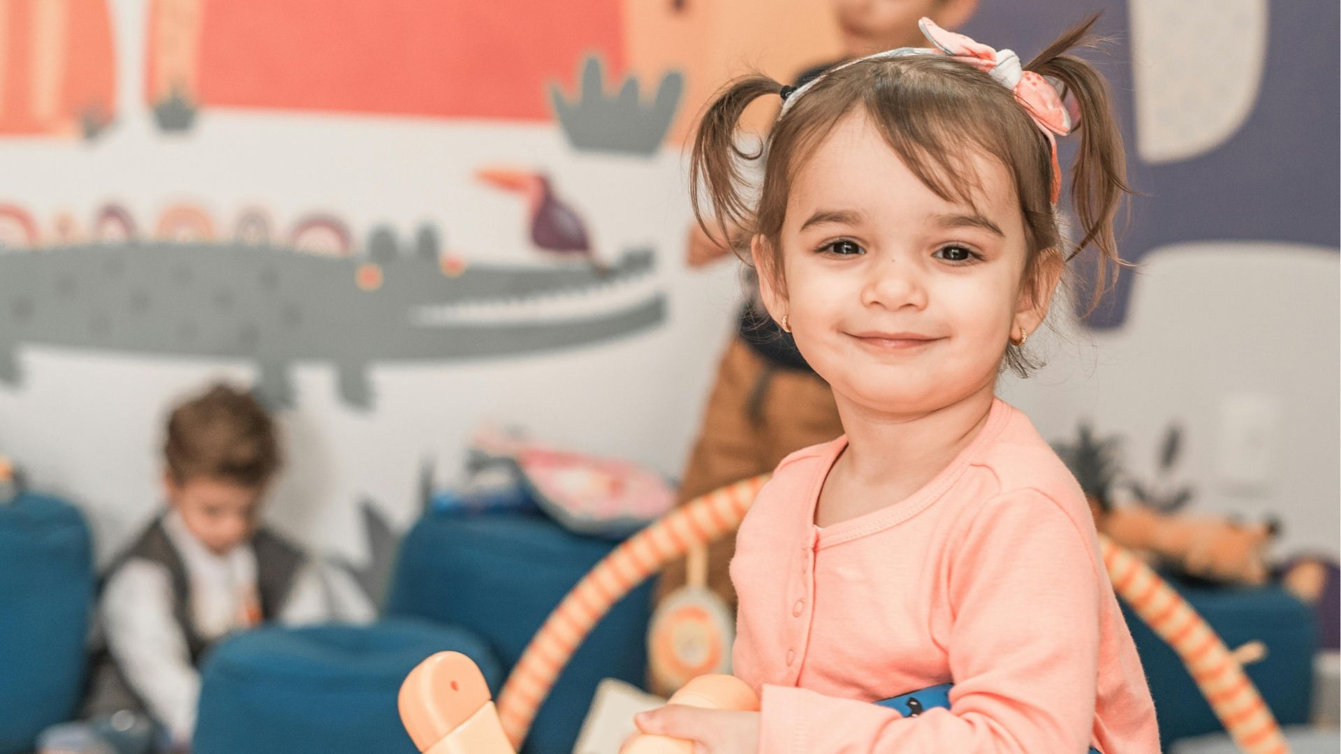 A smiling young girl in a childcare or early learning setting, holding a toy and standing in front of colourful, playful wall art. Other children are visible in the background, creating a happy and confident early childhood environment.