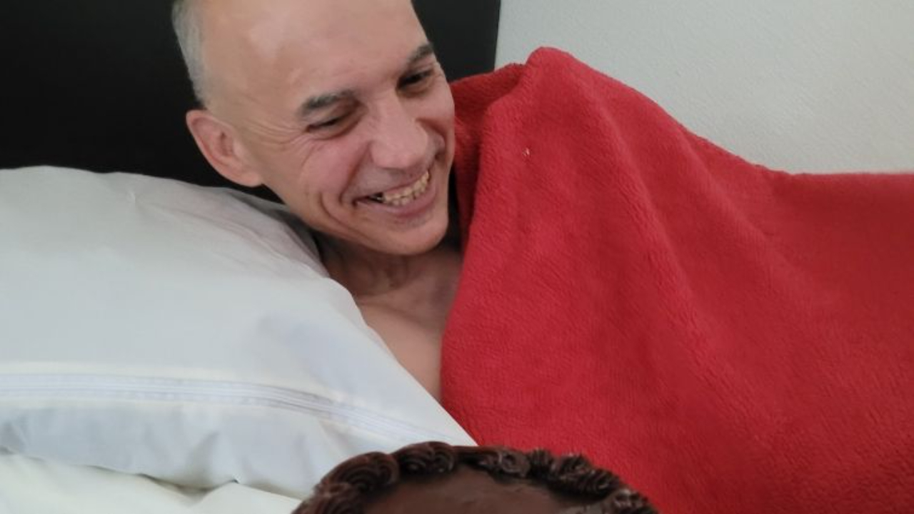 Hispanic man lays in bed covered with blanket and smiles as he looks at his chocolate birthday cake being held before him.