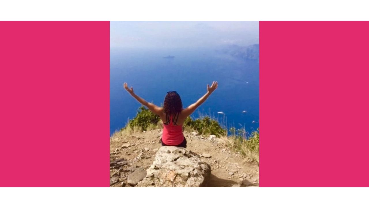 Jennifer sitting on a rock looking out at the ocean with her hands raised high