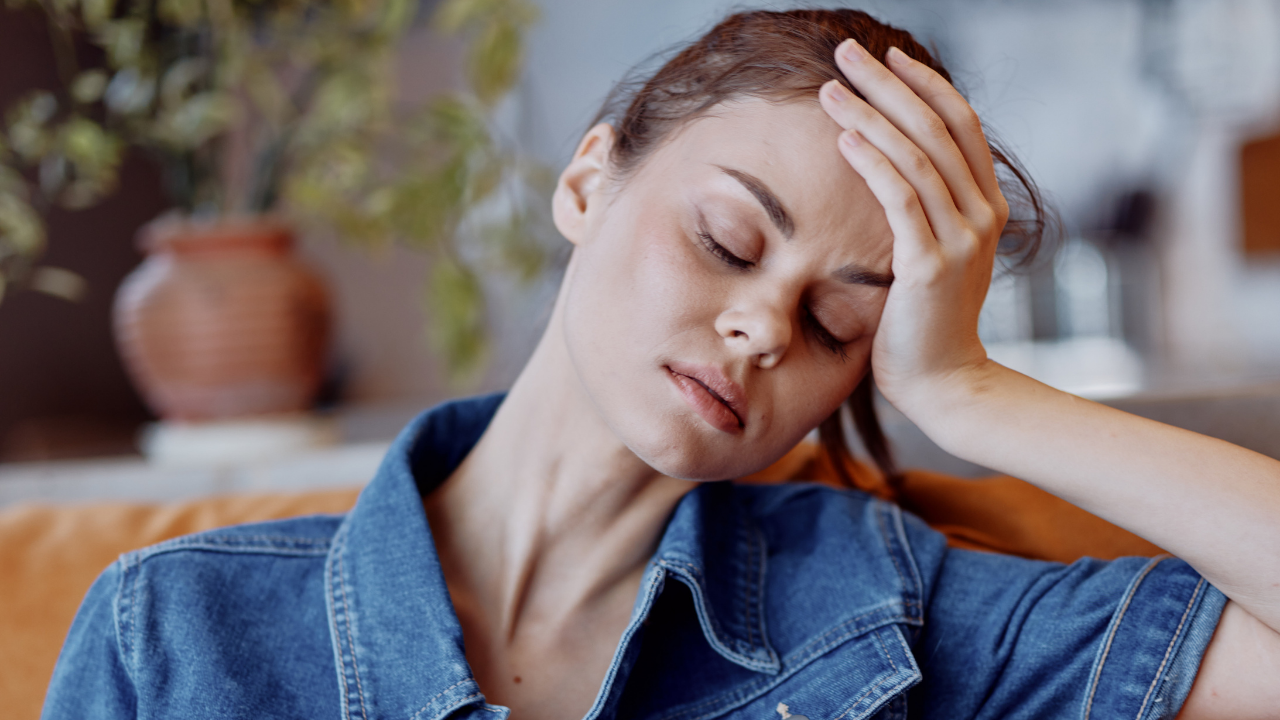 Woman sitting on a couch with her hand pressed to her forehead, eyes closed, looking stressed and overwhelmed, illustrating why stress feels different for women
