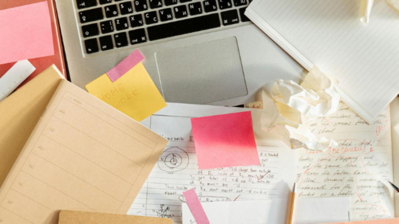 A cluttered desk with a laptop, notebooks, papers, sticky notes, and pens scattered across it.