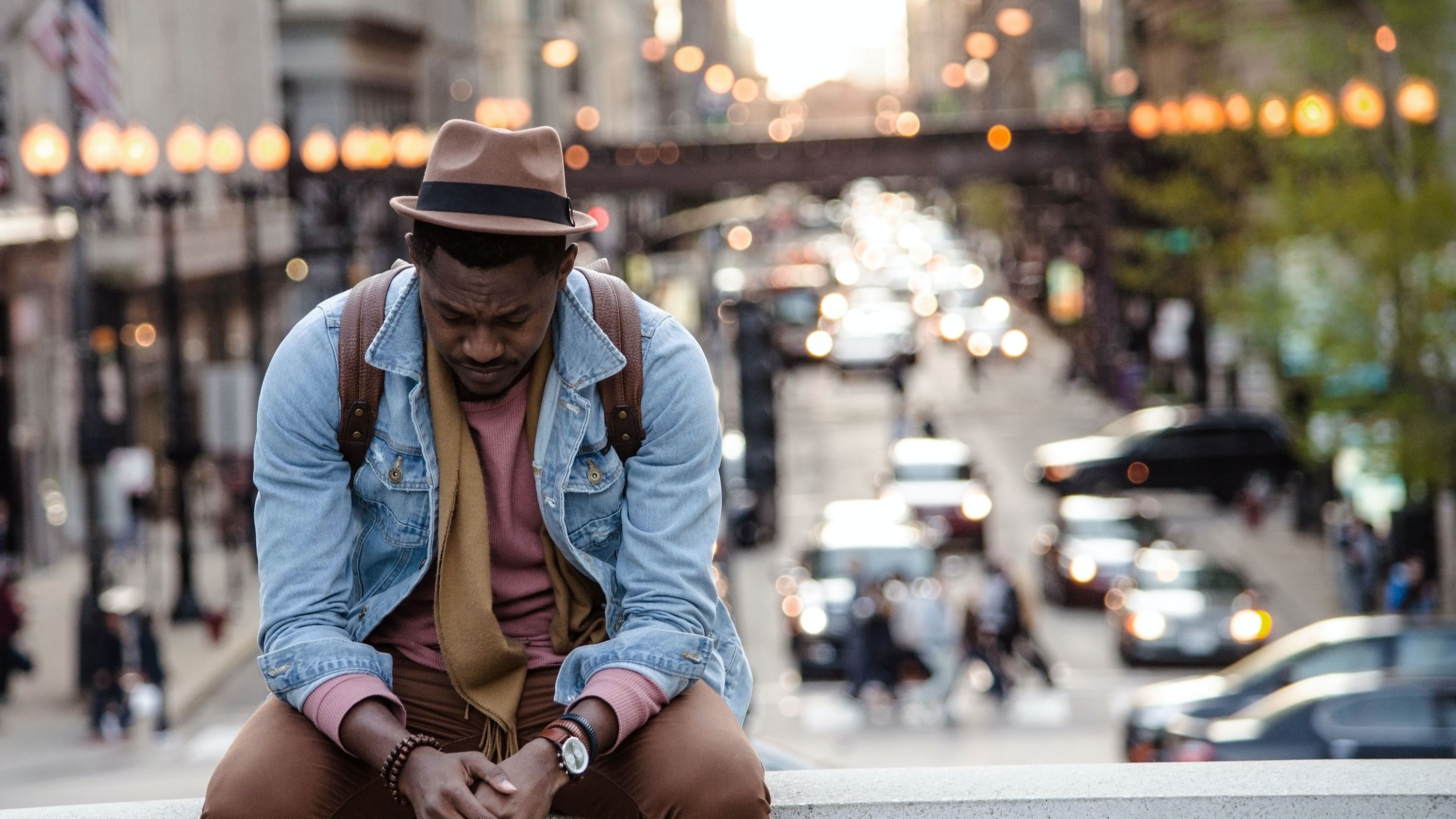 A man wearing a hat, denim shirt, and brown pants sits on a ledge facing the camera, with a city street behind him.