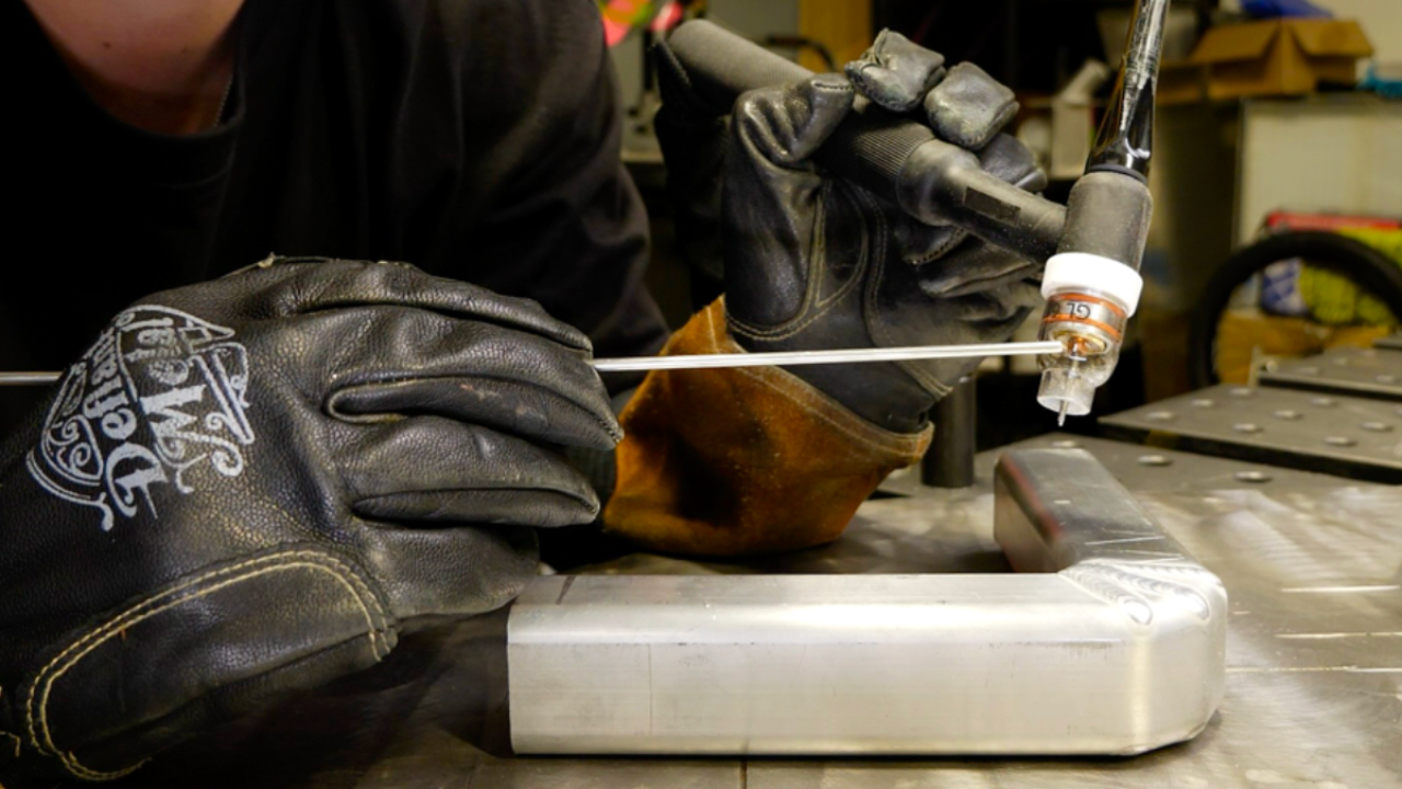 Close-up of a gloved hand demonstrating the proper TIG torch grip technique, showing Filler rod tips 