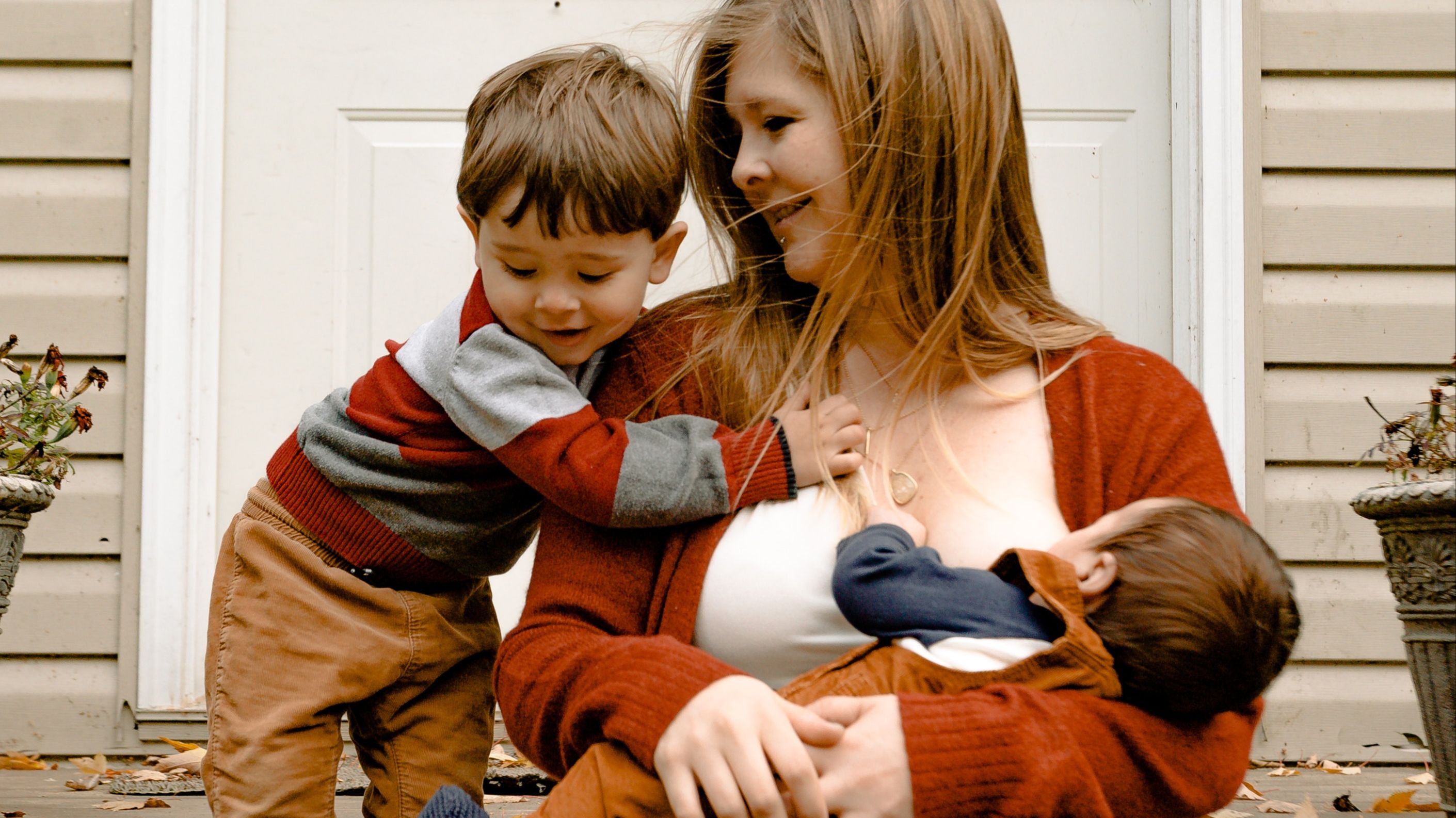 A white woman in a burnt orange sweater holding an infant nursing while looking at their toddler