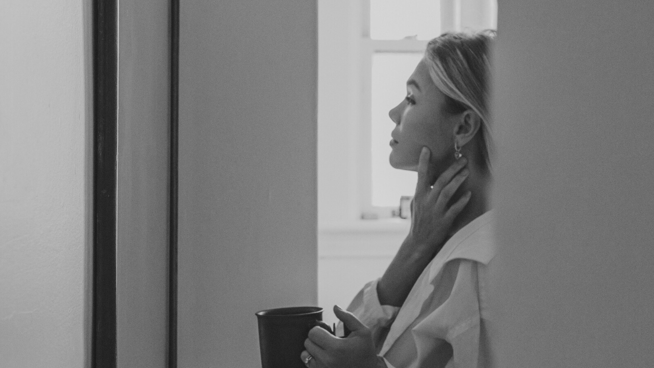 Woman standing by a window with a cup in hand, appearing lost in thought to symbolize self-reflection in relationships