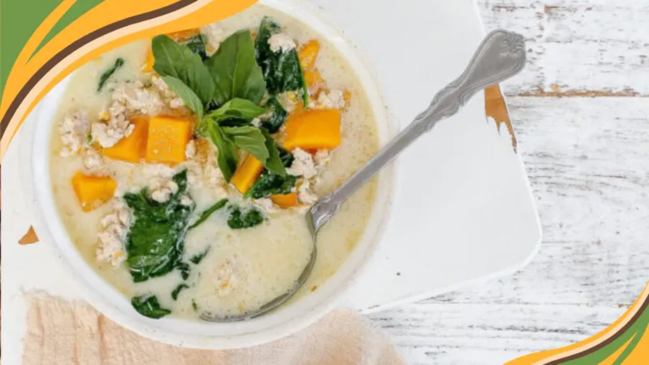 A white bowl of creamy soup topped with fresh basil, orange squash cubes, spinach, and shredded chicken, with a silver spoon resting inside, on a white wooden table.