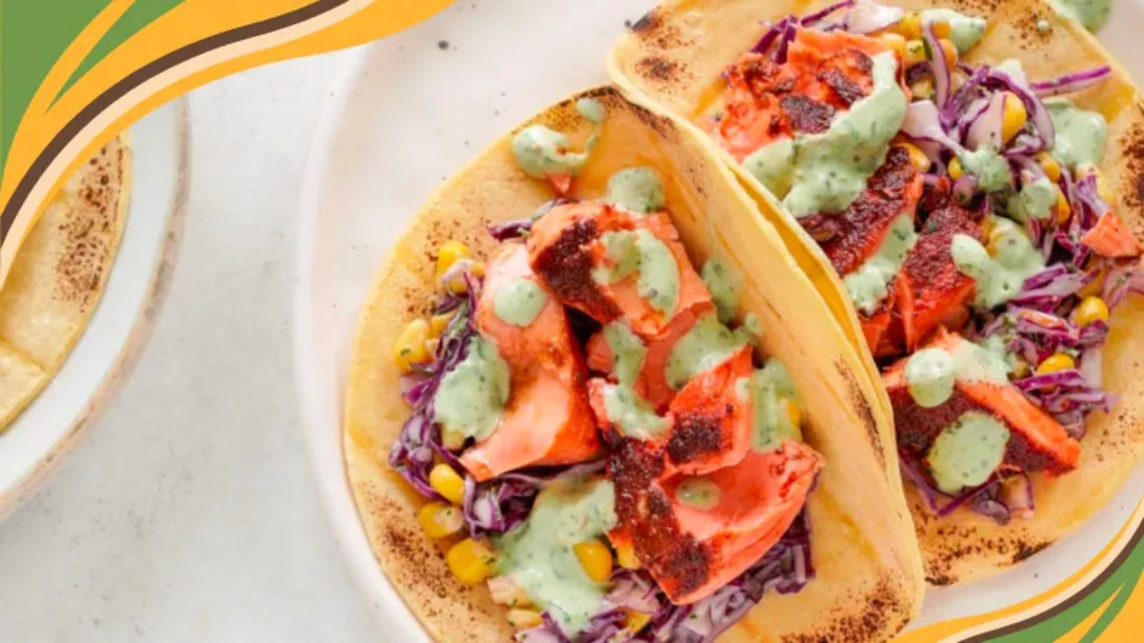 Two corn tortillas filled with flaky, seasoned salmon, shredded purple cabbage, and yellow corn, drizzled generously with creamy cilantro lime dressing. The tacos rest on a white plate with char marks visible on the tortillas, showcasing vibrant colors and fresh ingredients in a bright, appetizing presentation.
