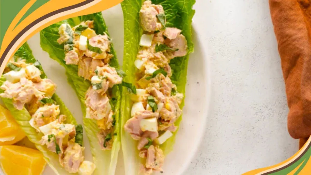 Four romaine lettuce leaves filled with chopped egg and tuna salad, garnished with fresh herbs, arranged on a white plate with lemon wedges. Extra lettuce leaves and a rust-colored napkin rest nearby on a light stone surface. Bright, fresh, and inviting.