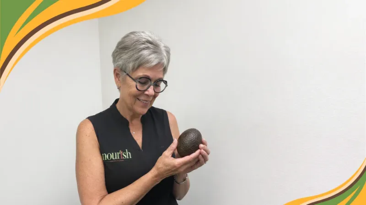 Woman over 50 smiling while holding a ripe avocado indoors, representing real food, nourishment, and the connection between avocados and better sleep for sustainable weight loss.