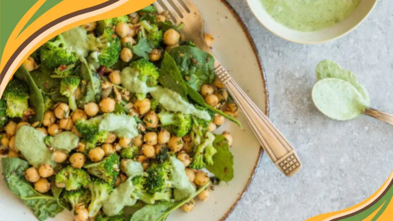 A shallow beige plate filled with a broccoli and chickpea salad on baby greens, drizzled with creamy green goddess dressing. A vintage silver fork rests on the rim. Right edge features curved green and gold graphic accents. Rustic glaze on the plate shows a thin brown ring. Dressing pools between tender florets and chickpeas. Lime nearby.