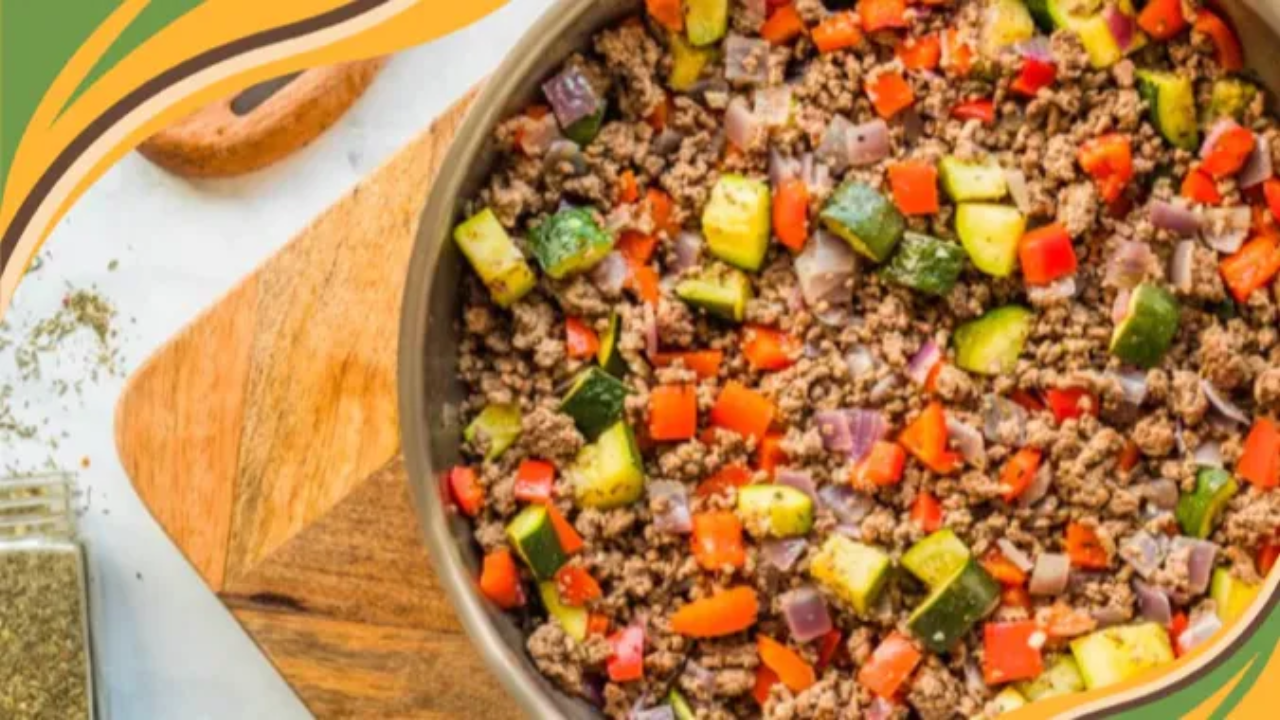A large silver skillet filled with ground beef, chopped red bell pepper, zucchini, and red onion, sitting on a wooden cutting board with a checkered tea towel and a jar of dried herbs nearby. A slotted wooden spoon, stacked plates, and serving utensils rest in the background. The scene feels fresh, vibrant, and homemade.