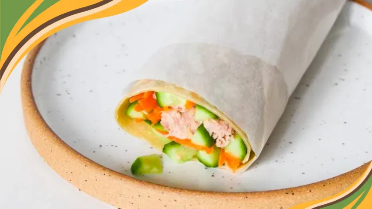 A spicy tuna wrap sliced in half, showing layers of shredded carrot, cucumber, and creamy tuna in a brown rice tortilla, served on a white plate with a side of chili flakes.