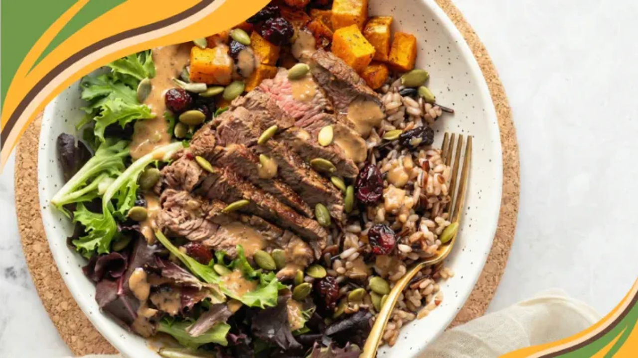 Overhead view of a composed salad bowl with sliced medium-rare steak over mixed greens, wild rice, roasted butternut squash, dried cranberries, and pumpkin seeds, drizzled with creamy dressing, with green and yellow decorative graphics in the top left corner.