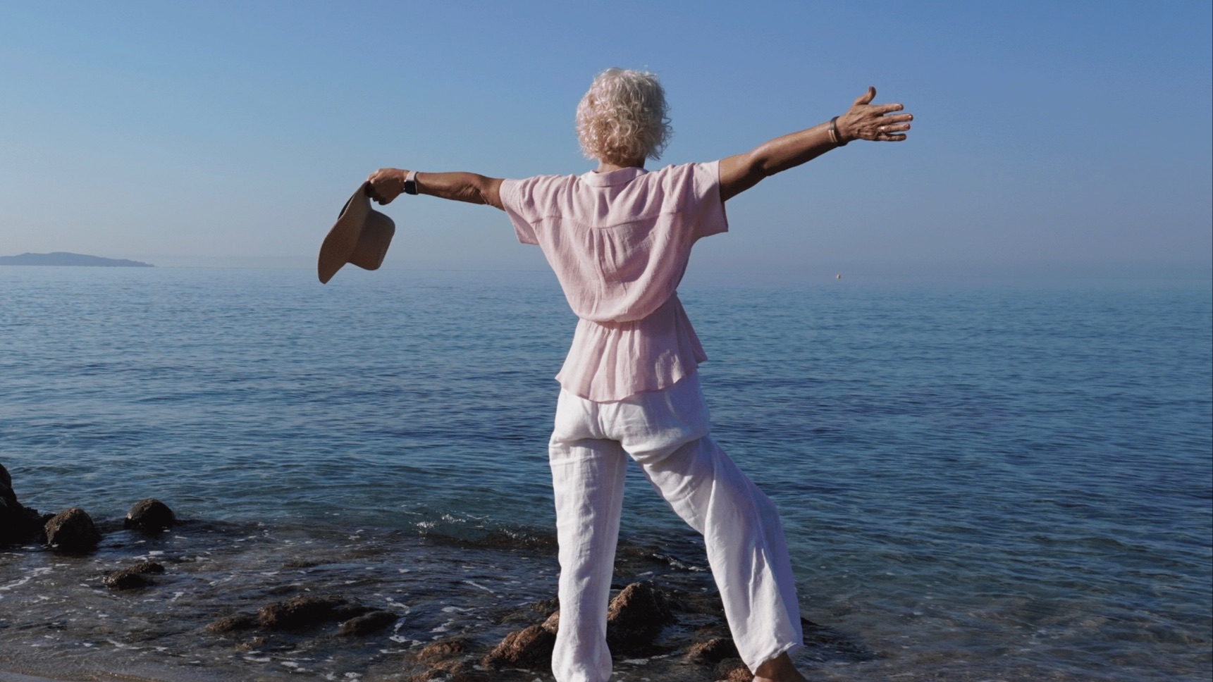 How to find yourself in menopause specialist, Julie Ann Garrido, stands on rock by ocean with arms wide holding a straw hat 