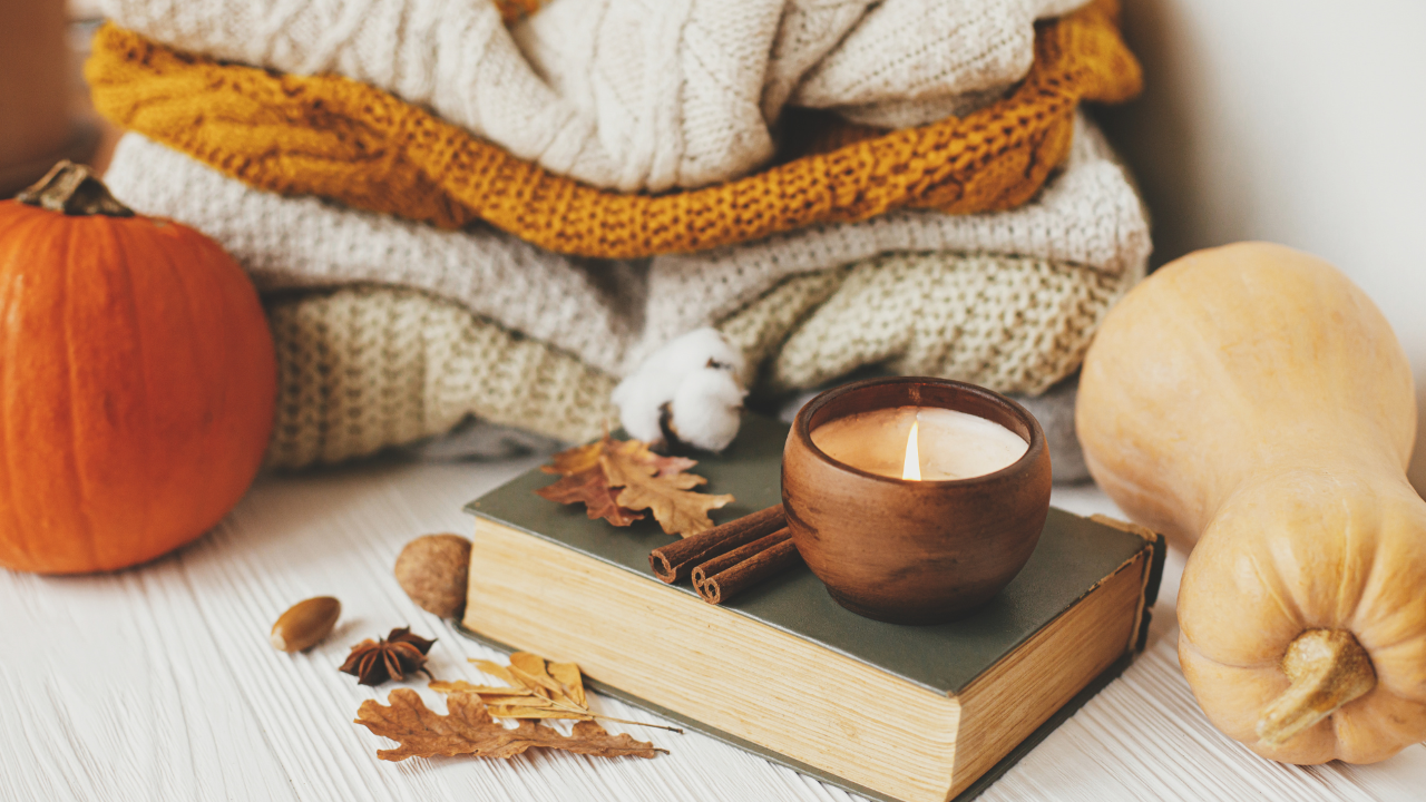 A stack of kit sweaters on a table top with a book, candle, fall leaves, and two gourds